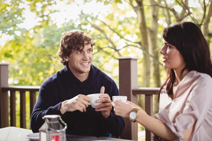 A man and a woman talking over coffee on a terrace among trees